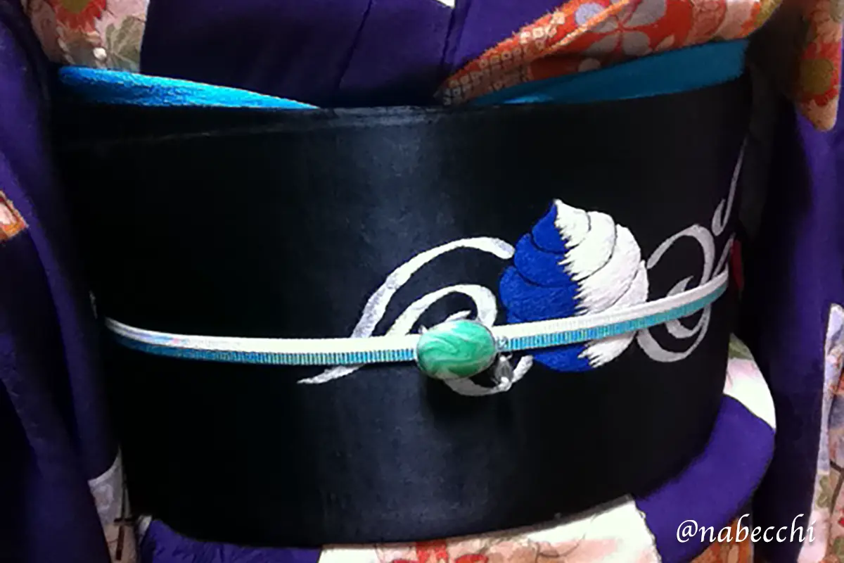 黒繻子帯の前面クローズアップ。青と白の宝珠刺繍とマーブル模様の帯留めでコーディネート。