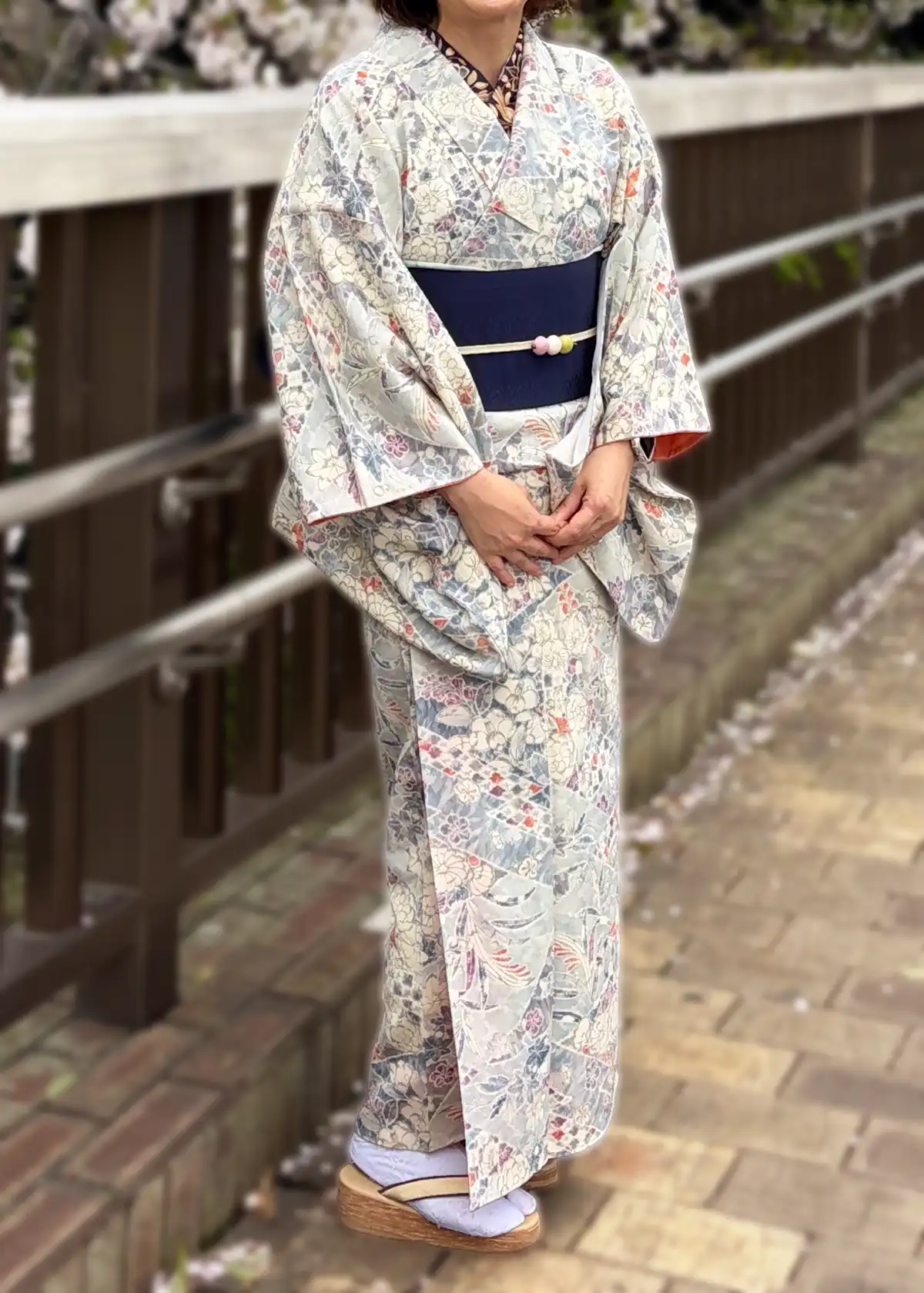 桜が満開の橋の上で、淡い色調の縮緬小紋と濃紺の博多帯、濃紺の刺繍半衿を合わせた全身コーディネート